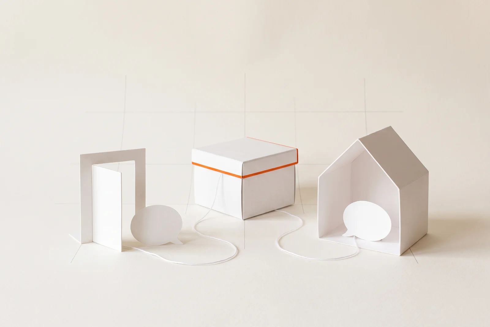 Zwei weiße Sprechblasen aus Papier, eine vor einer offenen Tür aus Karton, eine im Inneren eines Papierhauses, beide mit einem dünnen weißen Faden an dieselbe kleine Archivbox gebunden, deren Deckelkante orange ist